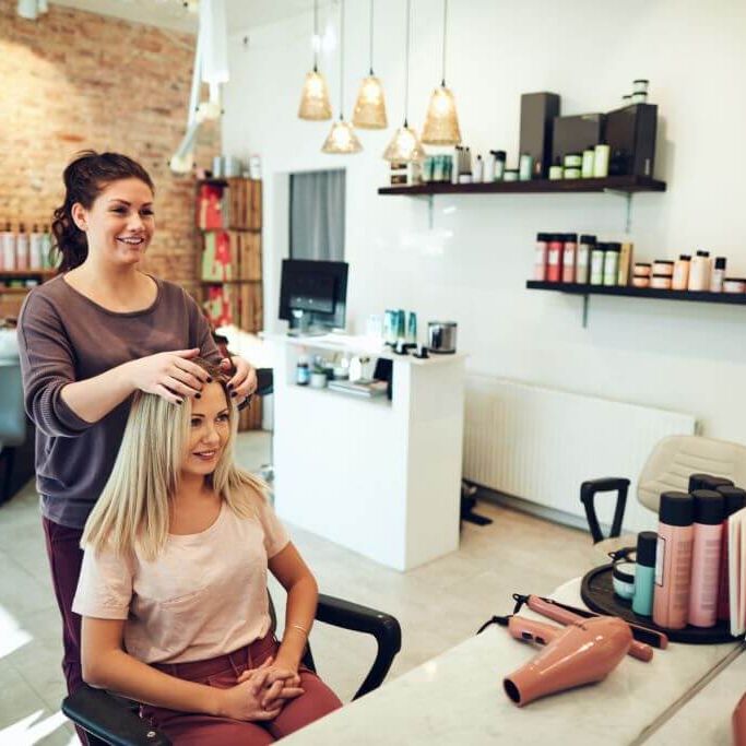 Young woman talking with her hairstylist during a salon appointment Young blonde woman smiling and looking at her reflection in a mirror while sitting in a salon chair during an appointment with her hairstylist